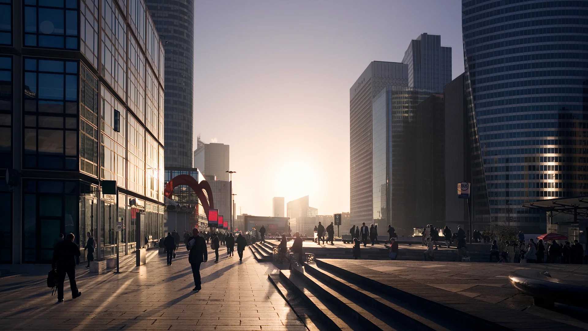 Silhouetted people walking on a wide plaza in La Défense, Paris, surrounded by modern high-rise office buildings under a sunset, showcasing a bustling real estate development business district.