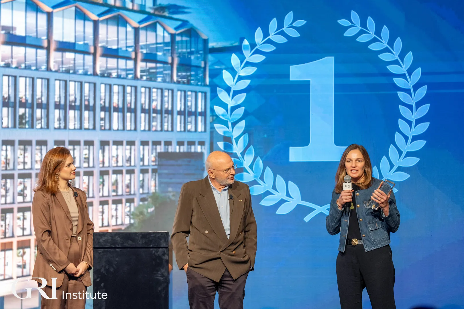 Three people on a stage at a real estate awards ceremony hosted by the GRI Institute, celebrating the Corporate Project of the Year winner.