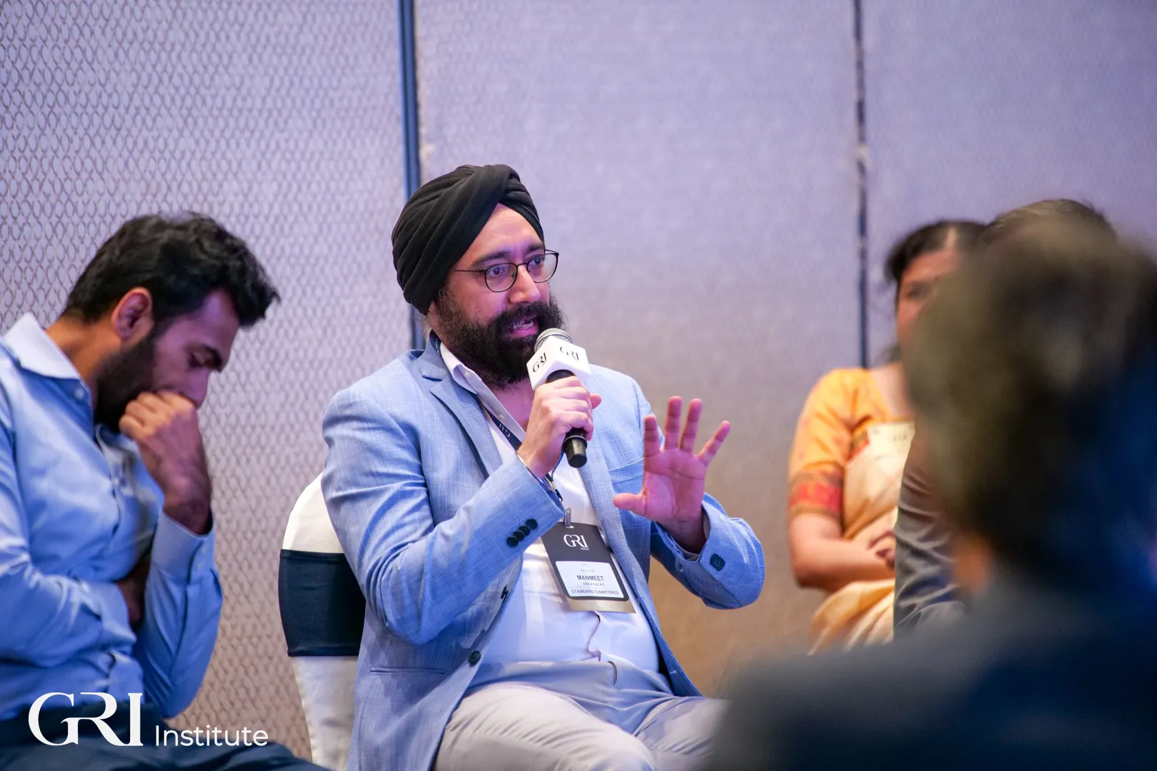 A male executive, holding a microphone, actively makes an intervention during a roundtable session, with other corporate leaders looking on at the GRI 2025 summit.