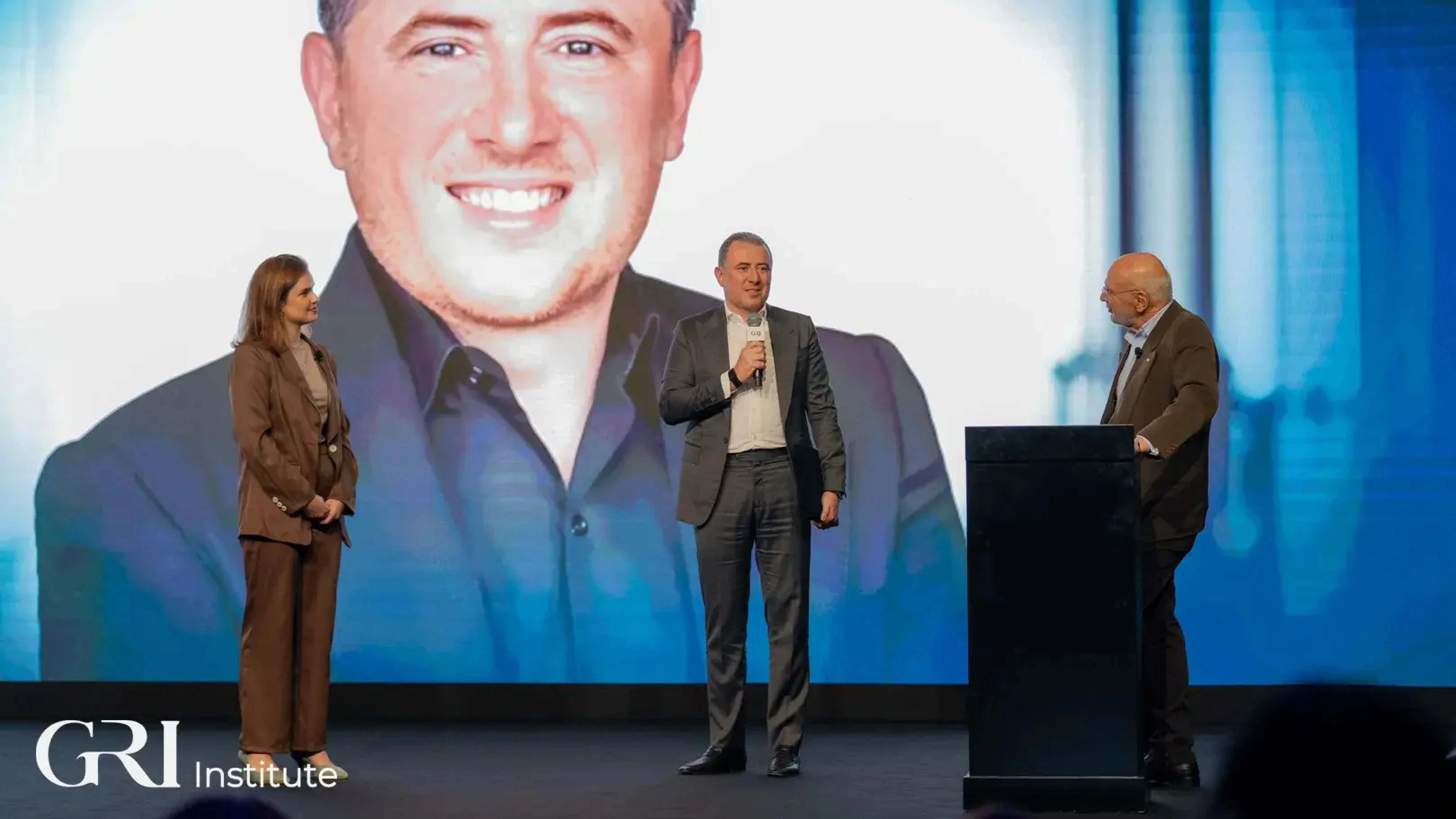 Three people on a stage at a real estate awards ceremony hosted by the GRI Institute, celebrating the Person of the Year winner.