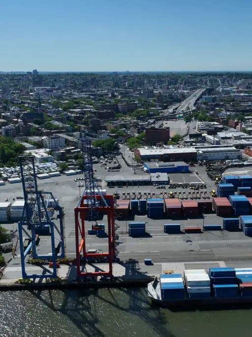 High-angle shot of blue and red gantry cranes at the Brooklyn port, with containers in the foreground and the Red Hook neighborhood in the back.